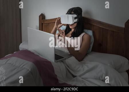 Contenu Femme dans VR lunettes assis sur le lit avec ordinateur portable et interagir avec la réalité virtuelle tout en jouant à un jeu vidéo Banque D'Images