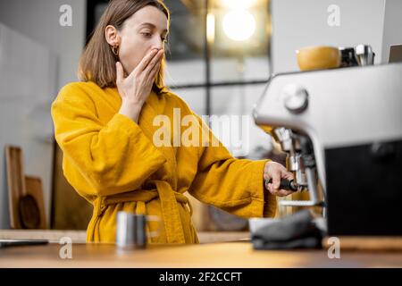 Jeune femme en jaune peignoir nawns en attendant le café machine à café professionnelle dans la cuisine le matin à la maison Banque D'Images
