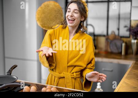 Une femme heureuse prépare des crêpes pour le petit déjeuner, en s'amusant à jeter des crêpes frites dans la cuisine. Bonjour et cuisine à la maison concept Banque D'Images