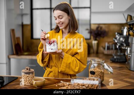 La jeune femme en peignoir jaune prend un petit déjeuner sain avec des céréales yaourt et baies dans le bol de la cuisine à à la maison le matin Banque D'Images