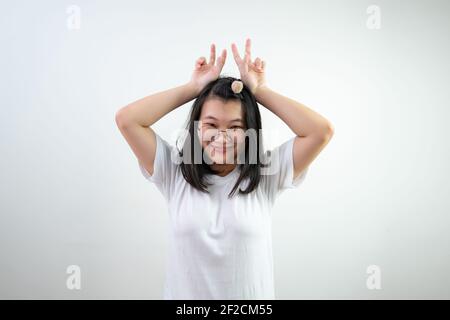 Lunettes asiatiques la jeune femme élève le signe de victoire par deux doigts comme les oreilles de lapin sur fond blanc. Banque D'Images