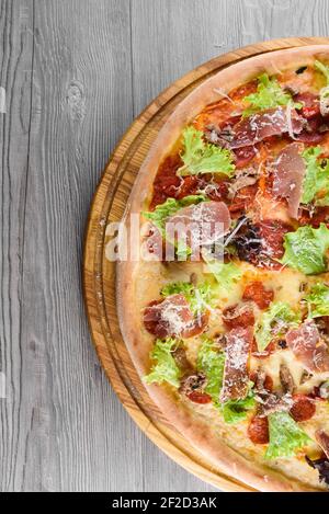 Délicieuse pizza italienne fraîche avec jambon, salami, tomates, salade et parmesan sur une planche en bois sur une table en bois. Vue de dessus. Banque D'Images