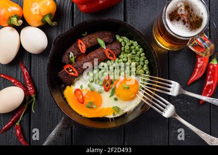 Repas aux protéines vives d'œufs biologiques frits, saucisses et petits pois servis pour deux avec bière et piment vif et poivrons belle sur fond noir au-dessus de vie Banque D'Images