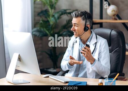 Un médecin sympathique, portant un blouse de laboratoire et un casque, parlant par appel vidéo avec un ordinateur et un patient, consultation en ligne. Concept d'aide médicale à distance Banque D'Images