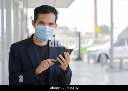 Homme d'affaires porter un masque médical utiliser smartphone regarder l'appareil photo extérieur, masque de protection du coronavirus covid19 Banque D'Images