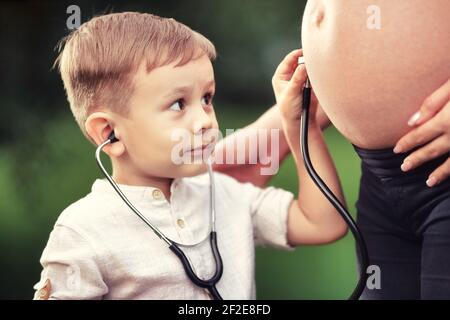 Un petit garçon écoute avec intérêt l'estomac d'une mère enceinte avec un stéthoscope. Banque D'Images