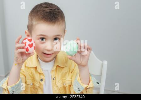 Adorable garçon présentant des œufs de Pâques peints. Un enfant blond a décoré des œufs de Pâques à la maison dans la cuisine. Un petit garçon adorable dans une chemise jaune sur un dos léger Banque D'Images