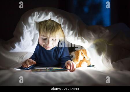 Adorable enfant blond, mignon garçon, lisant un petit livre avec un petit jouet à côté de lui sous la couette dans le lit Banque D'Images