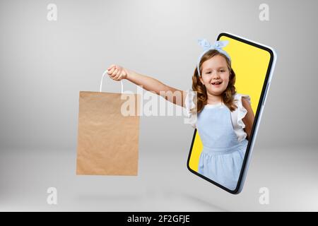 Une petite fille tient un sac cadeau dans sa main et sort du smartphone. Enfant en ligne sur l'écran d'un téléphone portable. Copier l'espace. Banque D'Images