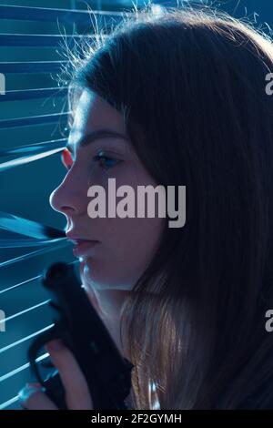 Une jeune femme avec une arme à la main regarde le fenêtre à travers les stores Banque D'Images