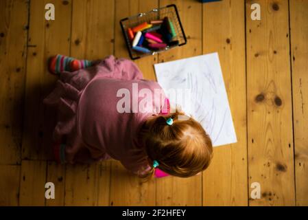 Une petite fille vêtue de rose et avec ses cheveux en queue de cochon se trouve sur un plancher en bois dessin sur papier blanc avec des crayons colorés. Banque D'Images