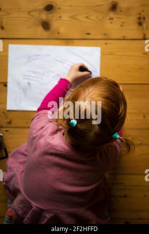 Une petite fille vêtue de rose et avec ses cheveux en queue de cochon se trouve sur un plancher en bois dessin sur papier blanc avec des crayons colorés. Banque D'Images