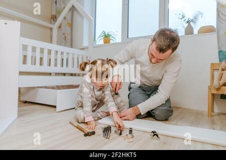 Une petite fille de 4 ans aide son père à assembler ou à fixer le tiroir du lit dans la chambre des enfants. Banque D'Images