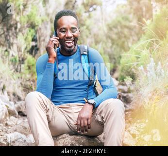 Un jeune homme ethnique africain parle par téléphone portable avec quelqu'un et riant avec joie lorsqu'il fait une randonnée dans la forêt tropicale. Bon peopl Banque D'Images