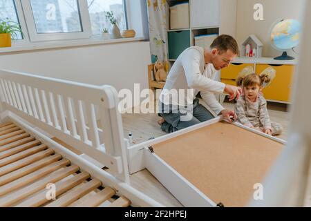 Une petite fille de 4 ans aide son père à assembler ou à fixer le tiroir du lit dans la chambre des enfants. Banque D'Images