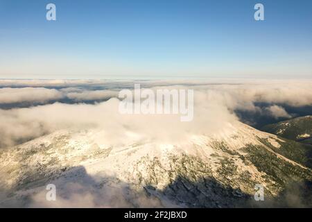 Vue aérienne depuis le dessus des nuages blancs bouffis couvrant les sommets enneigés des montagnes par beau temps ensoleillé. Banque D'Images