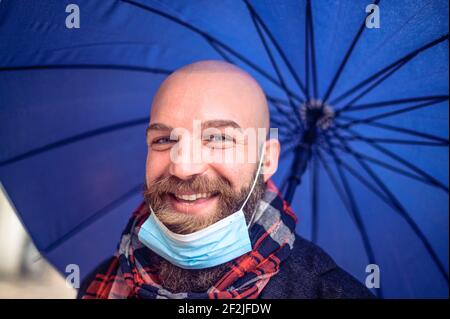 Taille basse chauve homme avec masque facial sous le port bleu de parapluie manteau d'hiver pour la pluie avec un visage heureux debout et sourire avec un sourire assuré Banque D'Images