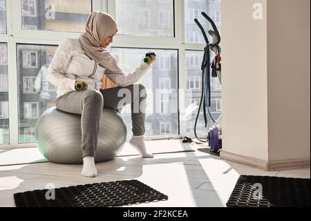 Belle femme musulmane arabe avec tête couverte dans le hijab assis sur une balle d'ajustement et pompant des biceps avec des haltères. Banque D'Images