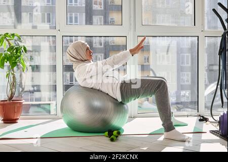 Charmante femme musulmane avec une tête couverte dans un hijab Exécute l'entraînement ABS avec les bras étirés sur un fitball dedans une salle de sport à la maison Banque D'Images