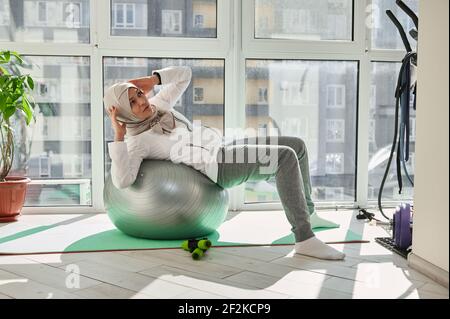 Charmante femme musulmane avec tête couverte dans hijab exécutant ABS faites de l'exercice sur un fitball à la salle de gym à la maison Banque D'Images