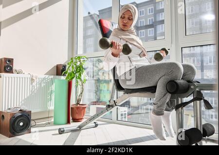 Jeune femme attirante dans hijab, exécute des exercices avec des haltères, assis sur un banc à la salle de gym à la maison. Culturisme, fitness, sport concept. Banque D'Images