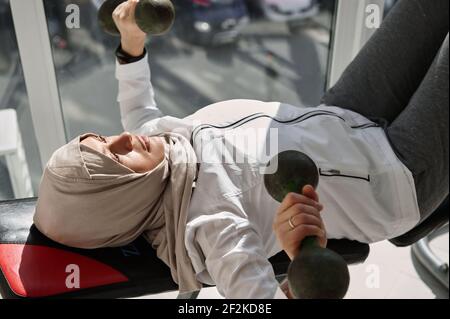 Vue en grand angle de la femme arabe musulmane déterminée avec la tête couvert faisant dumbbell banc de presse à la salle de gym à la maison contre le arrière-plan des grandes fenêtres Banque D'Images