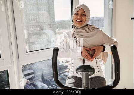 Femme arabe musulmane souriante avec tête couverte dans la démonstration de hijab une forme de coeur avec ses mains tout en faisant cardio Entraînement sur un vélo de spin à la homeA Banque D'Images