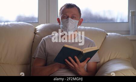 Jeune homme à la maison sur le canapé lisant un livre, auto-isolation, fond de fenêtre Banque D'Images