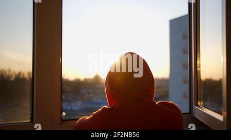 Un gars dans un sweat à capuche avec une capuche sur son la tête regarde le coucher du soleil en se tenant debout à l'air libre fenêtre Banque D'Images