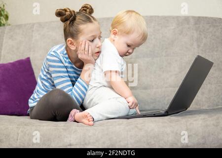 Petit garçon mignon et fille utilisant un ordinateur portable ensemble, regardant l'écran, regardant des dessins animés ou jouant en ligne, soeur et frère, frères et sœurs assis sur la comfor Banque D'Images