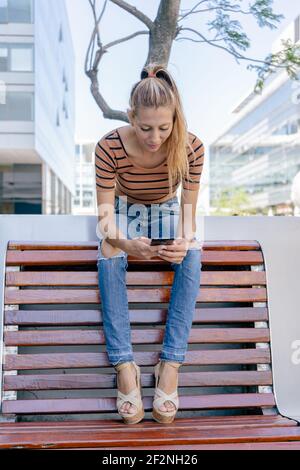 Belle femme latine vérifiant son téléphone portable confortablement installé sur un banc Banque D'Images