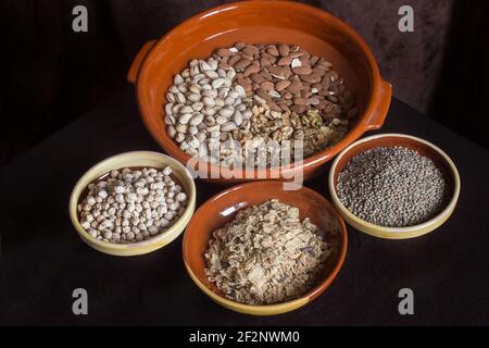 Un grand pot en argile avec une variété de fruits secs ainsi que de petites assiettes à soupe artisanales remplies de lentilles, de pois chiches et de flocons de riz grillés. Banque D'Images