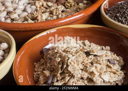 Gros plan d'une pile de riz rôti, de blé entier et de flocons d'orge avec des copeaux de chocolat entourés de bols en faïence remplis de noix sèches. Banque D'Images
