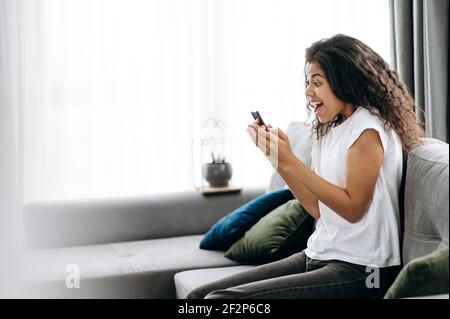 Une femme pigiste heureuse est surjoyeuse. Joyeuse jeune Afro-américaine femme assis sur le canapé, utiliser le smartphone, a reçu une bonne nouvelle, regarde joyeusement l'écran du téléphone Banque D'Images