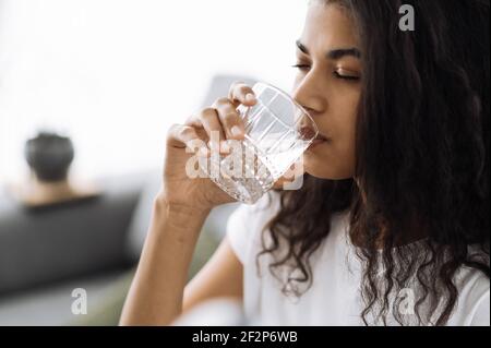 Un mode de vie sain. La jeune femme afro-américaine boit un verre d'eau tout en étant assise sur le canapé à la maison. Belle femme en bonne santé suivre un mode de vie sain Banque D'Images