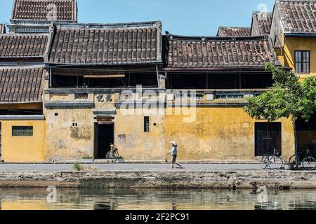 Femme vietnamienne portant un chapeau traditionnel non à la marche en face d'une maison de couleur pastel jaune classique avec des murs altérés à Hoi an, Vietnam Banque D'Images