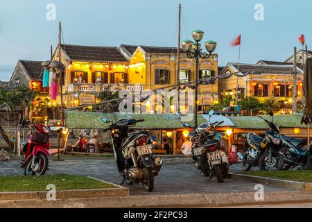 Une vue en soirée d'un restaurant de bateau de pêche amarré à la rivière Thu bon avec des bâtiments de restaurant éclairés à l'arrière, Hoi an, Vietnam Banque D'Images