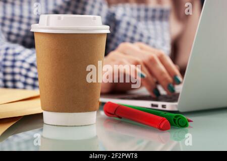 Une jeune femme boit du café dans une tasse jetable au bureau tout en travaillant avec un ordinateur. Gobelet en papier brun avec couvercle en plastique blanc Banque D'Images