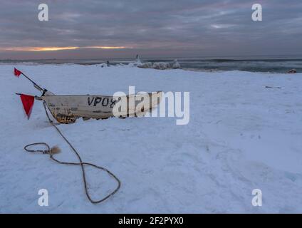 un bateau blanc est tiré sur la rive de la mer en hiver, la mer est couverte de neige mais il y a de la glace sous elle et il y a un coucher de soleil sur le ho Banque D'Images