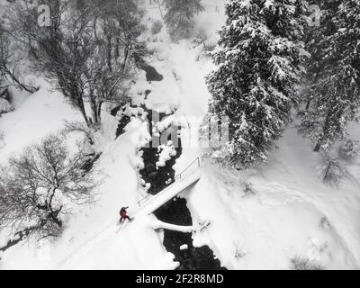 Vue aérienne par drone de la femme avec chaussures de spectacle traversant le pont à travers la rivière dans la forêt enneigée d'hiver à Almaty, Kazakhstan Banque D'Images