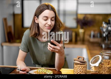 Femme en t-shirt vert décontracté prenant un petit déjeuner et bavardant sur un smartphone pendant une heure du matin dans la cuisine à la maison Banque D'Images