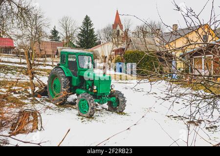 Maison typique en bois dans la campagne avec vieux tracteur vert Belarus dans le jardin en hiver, avec neige. La Biélorussie est une série de tracteurs à quatre roues Banque D'Images