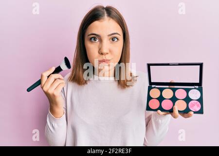 Jeune belle femme tenant la brosse de maquillage et rougir puffing joues avec le visage drôle. Bouche gonflée avec de l'air, attrapant l'air. Banque D'Images