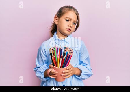 Petite belle fille tenant des crayons de couleur dans le visage de choc, regardant sceptique et sarcastique, surpris par la bouche ouverte Banque D'Images