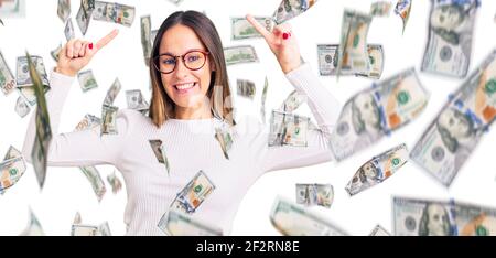 Belle brunette jeune femme portant un chandail blanc décontracté et des lunettes souriant émerveillé et surpris et pointant vers le haut avec les doigts et les bras relevés. Banque D'Images