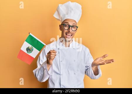 Homme chauve avec barbe portant un tablier de cuisinier professionnel tenant le mexique drapeau célébrant la réussite avec un sourire heureux et l'expression du gagnant avec élevé han Banque D'Images