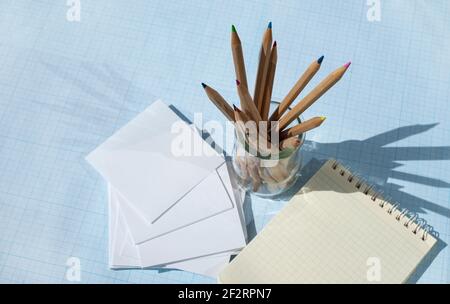 Crayons en bois avec fils multicolores dans un bécher en verre. Ombres nettes des crayons. Feuilles pour écrire et un bloc-notes carré. Banque D'Images