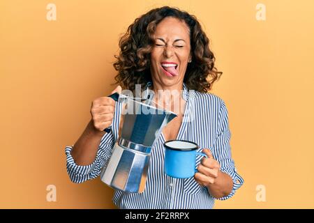 Femme hispanique d'âge moyen buvant le café italien collant langue dehors heureux avec l'expression drôle. Banque D'Images