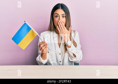 Jeune femme hispanique portant le drapeau ukrainien assis sur la table couvrant la bouche avec la main, choquée et peur de l'erreur. Expression surprise Banque D'Images