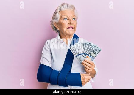 Femme sénior à cheveux gris portant le bras sur une élingue tenant des billets de banque en dollars américains souriant regardant sur le côté et regardant loin de penser. Banque D'Images